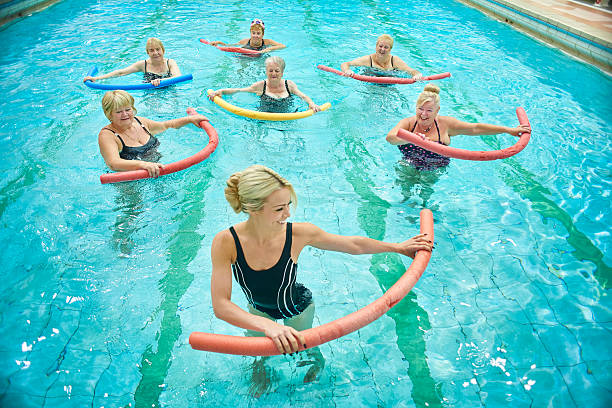 senior women led by female instructor at water aerobics A group of senior ladies take part in a water aerobics session. They are using pool noodles and are being led by a young female instructor. They are all laughing and smiling water aerobics stock pictures, royalty-free photos & images