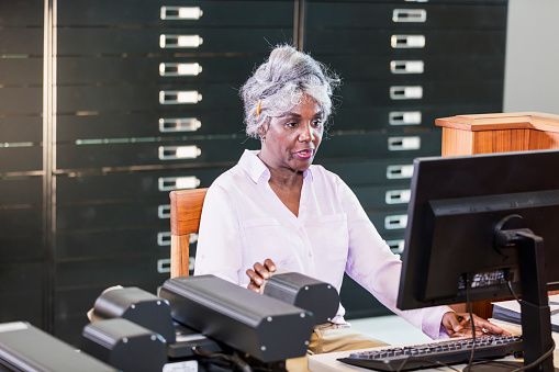 A senior African-American woman in the library, using a computer to review historical documents stored on microfiche.