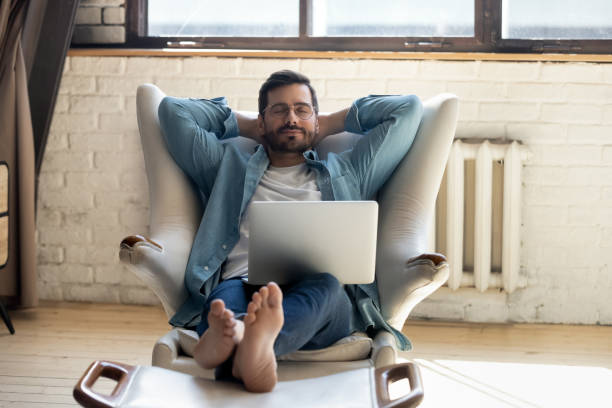 Serene barefoot guy resting, daydreaming at home. Front view peaceful young man in eyewear crossed hands behind head, fallen asleep on cozy armchair with footstool during remote workday with computer. Serene barefoot guy resting, daydreaming at home. barefoot stock pictures, royalty-free photos & images