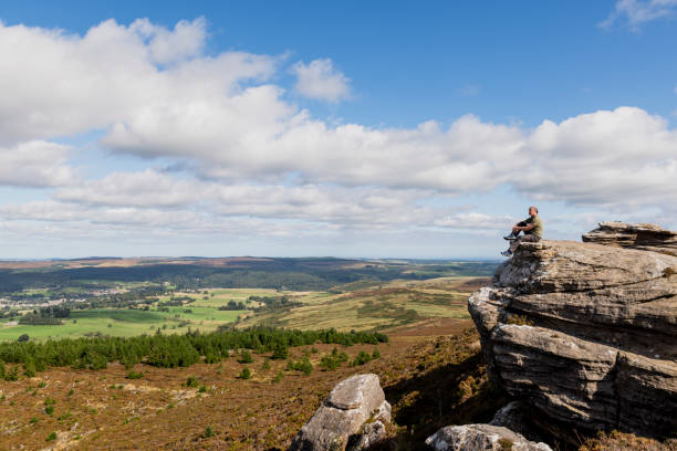 A Serene Individual A senior man sitting on a cliff edge and looking at the view while out on a hike in Rothbury, Northumberland. The beautiful green scenery is surrounding him. rothbury northumberland stock pictures, royalty-free photos & images