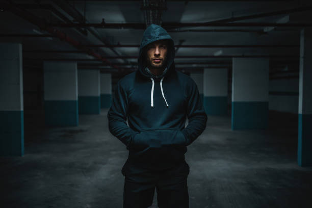 Serious attractive muscular caucasian sportsman in hoodie standing in underground garage with hands in pockets at night. Urban life concept. Serious attractive muscular caucasian sportsman in hoodie standing in underground garage with hands in pockets at night. Urban life concept. hooded shirt stock pictures, royalty-free photos & images