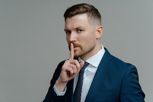 Private information. Young businessman in formal wear suit showing gesture of silence or shh sign, keeping finger near mouth and looking at camera while standing against grey studio background