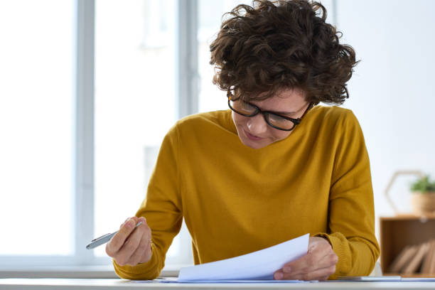 Serious busy young lady with short curly hair sitting at desk and filling document while preparing tax papers Busy lady filling document at workplace form filling stock pictures, royalty-free photos & images