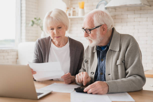 Serious caucasian old elderly senior couple grandparents family counting funds on calculator, doing paperwork, savings, paying domestic bills, mortgage loan, pension at home using laptop. stock photo