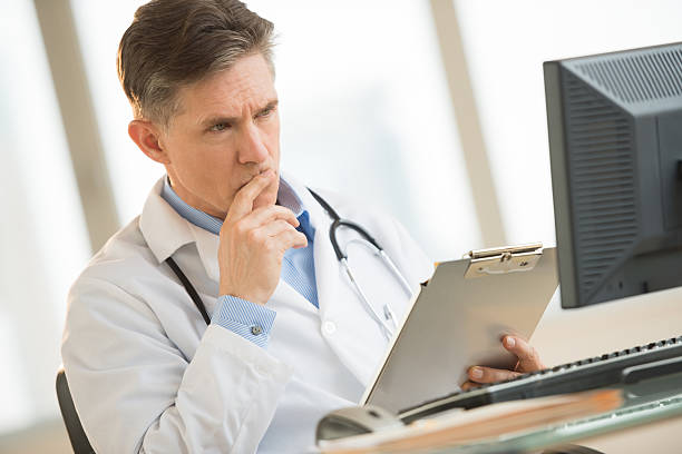 Serious Doctor Looking At Computer While Holding Clipboard In clinic stock photo
