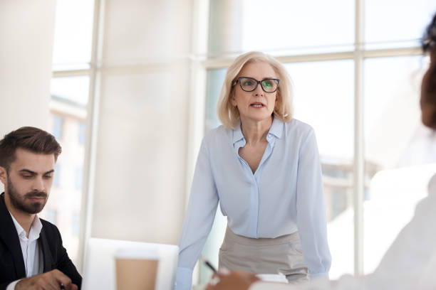 Serious mature businesswoman talk having discussion during briefing Serious mature businesswoman stand talk with employees at office meeting, middle aged female boss discuss project or strategy with workers at office briefing, confident woman negotiate with colleagues midsection stock pictures, royalty-free photos & images