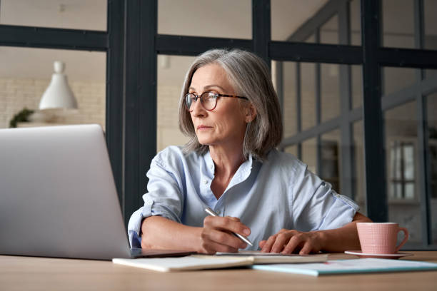 自宅やオフィスで仕事ノートパソコンでトレーニングウェビナーを見て深刻な成熟した成人女性。テーブルに座っているコンピュータ技術を使用しながらメモを取る60年代の中年ビジネスウ� - 定年　女性 ストックフォトと画像