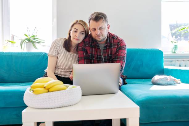 Serious middle-aged couple husband and wife with laptop sitting at home on couch stock photo