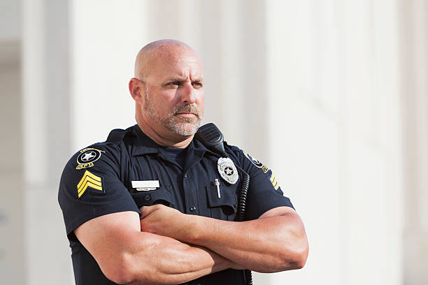 Close up of a police officer in uniform, standing with a stern look on his face, and arms crossed, watching something in the distance. He is in his late 40s, with a bald head and gray beard, mustache. and muscular build.