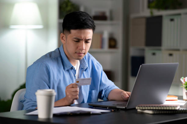 Serious young Asian man, frowning and worried while using and looking at credit card, doing online shopping. stock photo