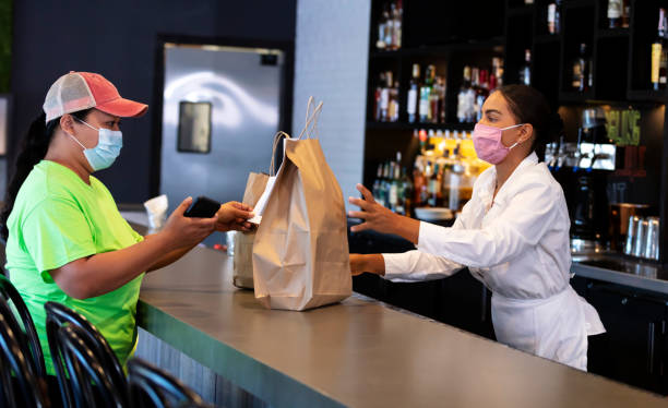 Server giving takeout food order to delivery person, masked A restaurant worker giving paper shopping bags to a delivery person who is picking up a takeout food order. She is looking at her mobile phone to verify the order. Both are mid adult mixed race women in their 30s. The waitress is African-American, Native American and Caucasian. The delivery person is Filipino and Cambodian. They are wearing protective face masks, working during the COVID-19 pandemic, trying to prevent the spread of coronavirus. restaurant delivery app stock pictures, royalty-free photos & images