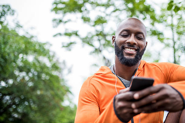 Setting the right mood for training African american man sitting outdoors before his training. He is holding mobile phone and looking at it. active lifestyle stock pictures, royalty-free photos & images