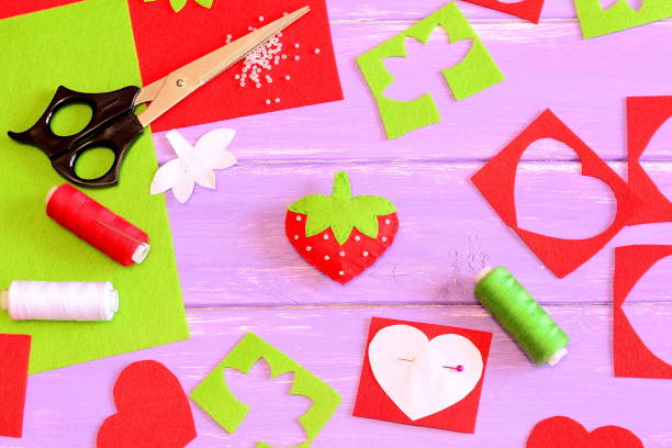 Sewing toy, tools and materials. Felt strawberry toy food, scissors, red green felt sheets and scraps, thread, sewing items, paper pattern on lilac wooden background. Abstract strawberry craft background. Top view stock photo