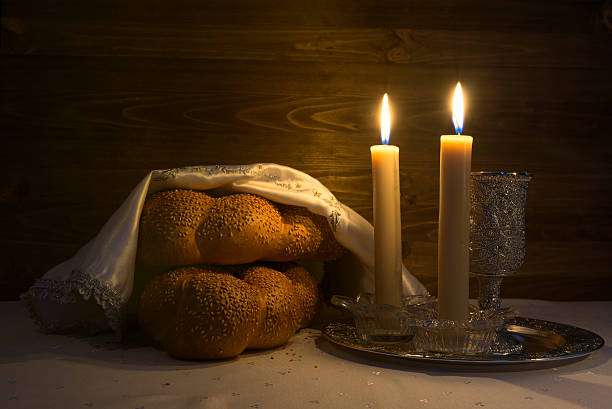 Softly and lowly lit photo of two loaves of challah, two shabbat candles, and a kiddush cup.