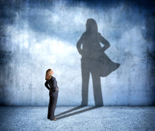 Shadow Of Woman Wearing A Hero's Cape A woman stands with her hands on her hips as she looks up at her shadow that is cast on the wall in front of her that shows her in a confident stance and wearing a cape. cape garment stock pictures, royalty-free photos & images