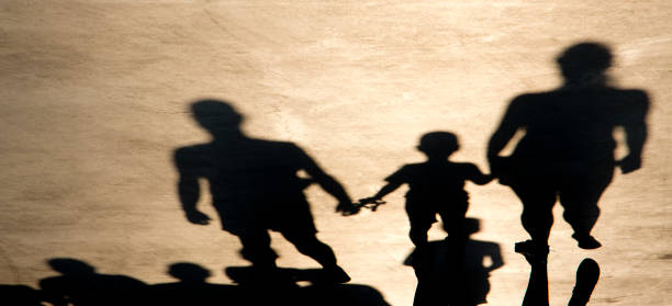 Shadow silhouette of parents and son holding hands while walking toward on summer road and many people behind Shadow silhouette of mother, father and son holding hands while walking toward on misty summer road and many people behind them, in sepia black and white approaching photos stock pictures, royalty-free photos & images