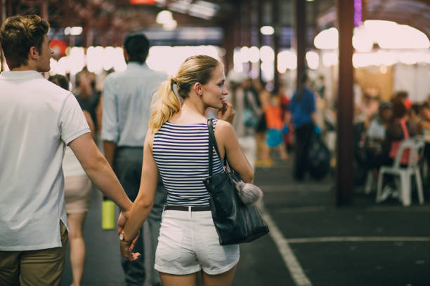 Shall I Buy It? Young couple are exploring Queen Victoria Market in Australia. They are holding hands and the woman is looking out oft he frame to something she is tempted to buy. queen victoria market stock pictures, royalty-free photos & images