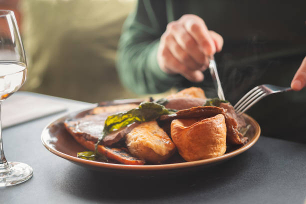Shallow focus on  yorkshire pudding as part of a meal of roast beef and vegetables. A mans hands can be seen using a knife and fork. A wine glass is on the table. stock photo