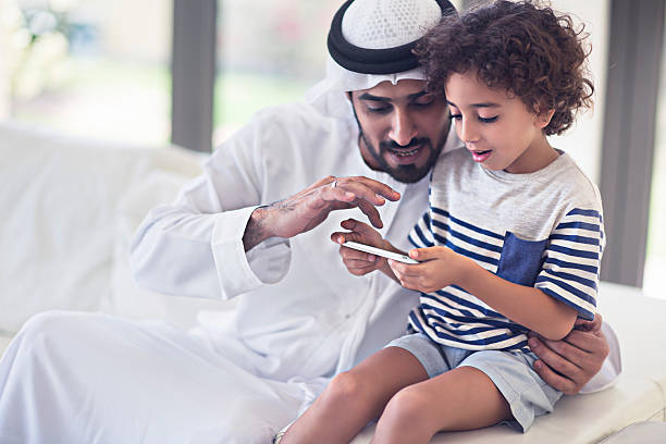 Sharings time with his son Middle Eastern father sharing time with his son at home, father is teaching how to use a mobile phone application,the father is wearing the traditional Dishdasha. arabia stock pictures, royalty-free photos & images