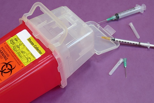 A red, plastic sharps container (with a biohazard symbol) with needles (syringes) on a purple background.