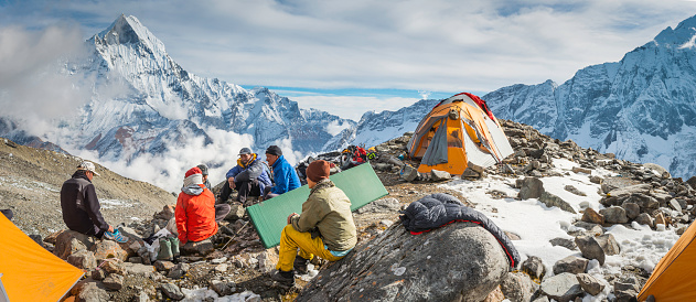 Deurali, Nepal - 2nd November 2012: Team of Sherpa mountaineers sitting on rocks amongst base camp tents overlooked by the iconic pyramid peak of Machapuchare (6993m) deep in the Himalayan mountain wilderness of the Annapurna Conservation Area, Nepal. Composite panoramic image created from five contemporaneous sequential photographs. 