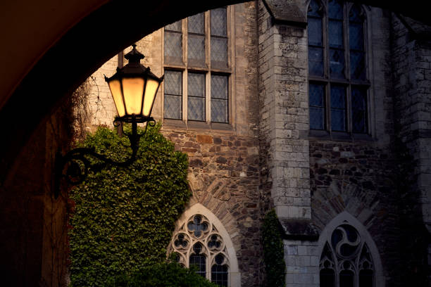 Shining yellow lantern in front of medieval sacral building with high windows and stone walls, view through dark vault stock photo