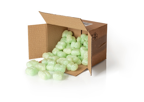 Shipping peanuts spilling out of the parcel box laying on its side, isolated on a white. Brown cardboard box with loose green foam fillers to protect fragile parcels in transit. Polystyrene cushioning pellets. Front view.