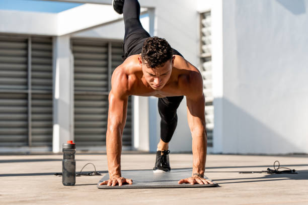 Shirtless muscular male athlete doing high plank leg lift exercise in the open air on building rooftop floor stock photo