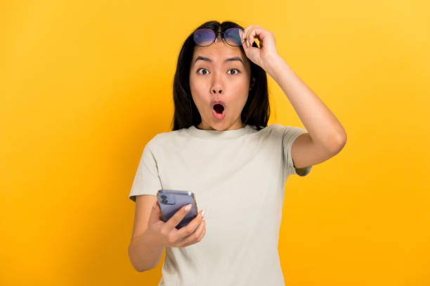 ショックを受けた驚いた中国の若いブルネットの女性は、携帯電話を手に持ち、カメラに驚いた様子で、眼鏡を外して口を開け、カジュアルなtシャツで孤立したオレンジ色の背景に立ってい - 驚き ストックフォトと画像
