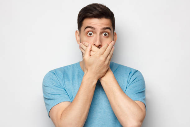 schockierter mann in blauem t-shirt gekleidet, bedeckt mund mit beiden händen, augen rund mit schock und angst, isoliert auf grauem hintergrund - mund stock-fotos und bilder