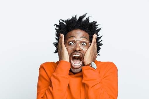 Terrified afro american young man wearing orange hoodie and glasses, looking away and screaming. Studio shot on grey background.