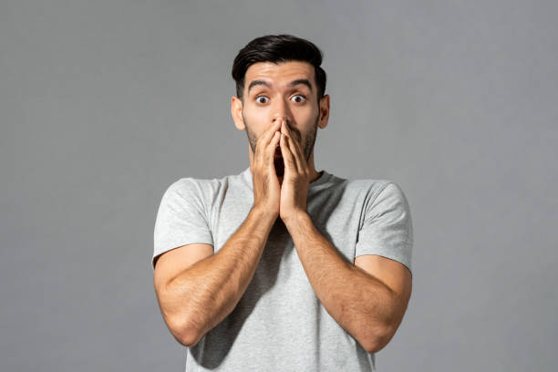 Shocked young Caucasian man with hands covering mouth in isolated light gray studio background stock photo