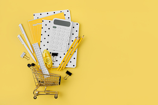 Shopping cart with School stationery supplies on yellow background. Concept back to school. Blank notebook with copy space