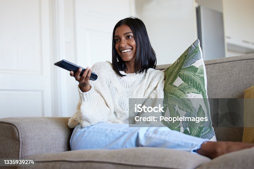 istock Shot of a beautiful young woman holding a remote control while sitting on the couch at home 1335939274
