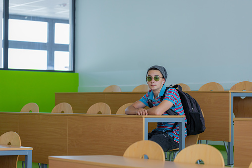 A photo of a cool young student sitting in the amphitheater with a backpack and headphones, wearing a cap and sunglasses, looking at the camera.