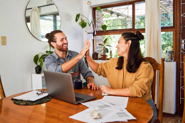 Shot of a happy young couple sharing a high five while using a laptop and checking paperwork at home stock photo