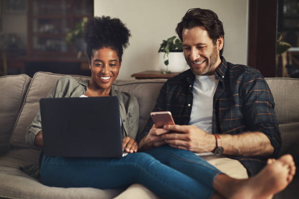 Shot of a happy young couple using a laptop and cellphone while relaxing on a couch ho Divided attention streaming service stock pictures, royalty-free photos & images