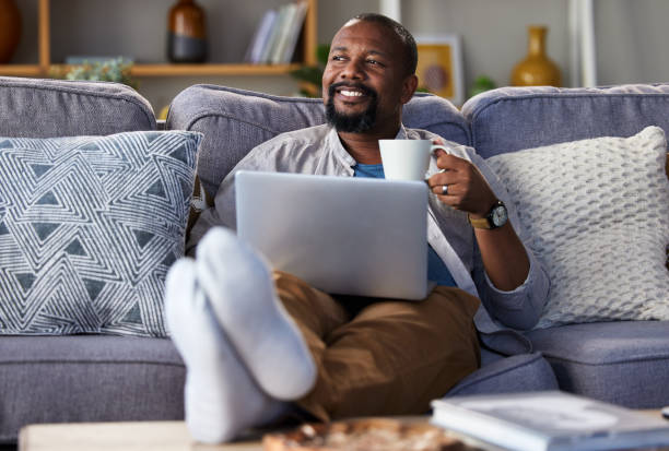 Shot of a mature man using a laptop and having coffee on the sofa at home Makes you think, doesn't it? electronic banking stock pictures, royalty-free photos & images