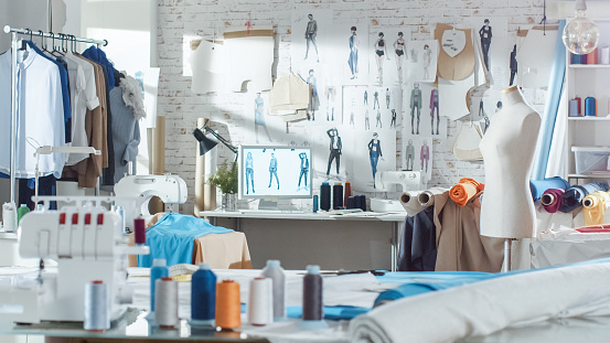 Shot of a Sunny Fashion Design Studio. We See Working Personal Computer, Hanging Clothes, Sewing Machine and Various Sewing Related Items on the Table, Mannequins Standing, Colorful Fabrics.