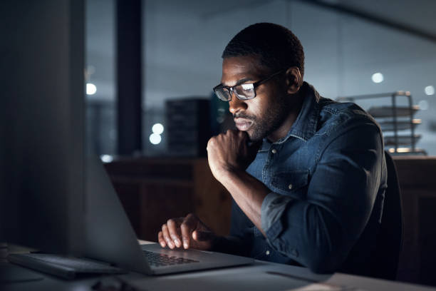 Shot of a young businessman using a laptop during a late night in a modern office stock photo