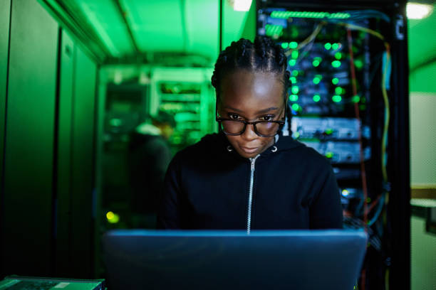 Shot of a young female computer programer using a laptop while standing in a dark server room stock photo