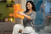 istock shot of a young women with teddy bear sitting on window sill at home, stock photo 1305075572