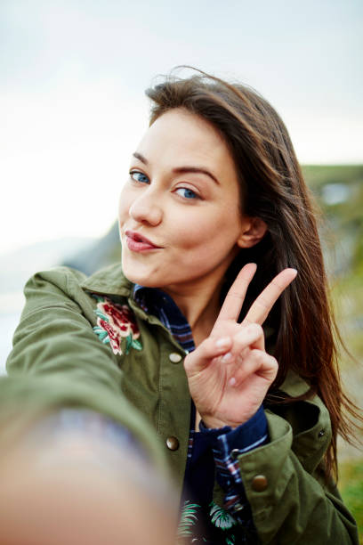 POV shot of beautiful woman posing while taking selfie against sky Portrait of beautiful woman puckering lips and gesturing peace sign while taking selfie on mountain. POV shot of young female tourist is against clear sky. She is wearing casuals. irish women stock pictures, royalty-free photos & images