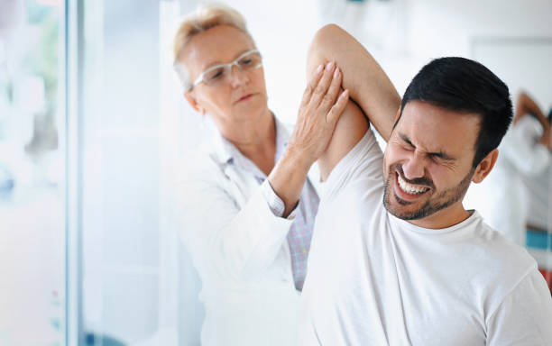 Shoulder problems. Closeup front angle view of a late 50's female doctor examining a male athlete with some shoulder pain. She's rotating and twisting his shoulder joint and trying to determine which tendons have been damaged. The patient has a painful grimace on his face. sports medicine stock pictures, royalty-free photos & images