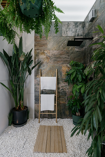 Shower cabin with wooden hanger for fresh and clean towel in modern apartment. Vertical shot of tropical bathroom with brown marble tiles on walls, pebble floor, and different green plants
