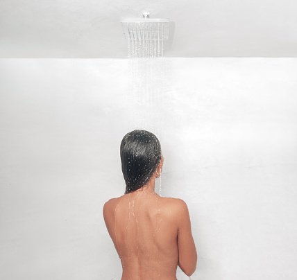 Shower lifestyle. Woman taking a hot bath showering washing her hair in luxury hotel bathroom with rainfall showerhead feature. View from behind.