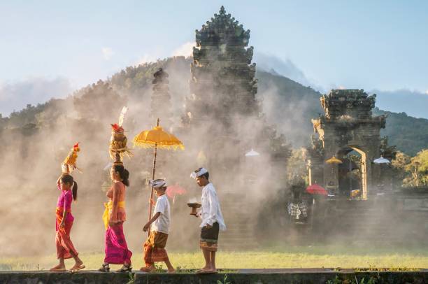 Showing traditional Balinese male and female ceremonial clothing and religious offerings as a mother and children walk to a Hindu temple (pura) in Bali. A mother and a teenaged girl are dressed in brightly colored sarongs, blouses, and sashes and are balancing tall fruit baskets on their heads. Two sons are dressed in sarongs and white shirts. The family is walking in front of an old stone temple building which has a smoky atmosphere. bali stock pictures, royalty-free photos & images