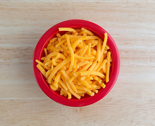 Shredded sharp cheddar cheese in red bowl Top view of a bowl of shredded sharp cheddar cheese on a wood table top illuminated with natural light. cheddar cheese stock pictures, royalty-free photos & images
