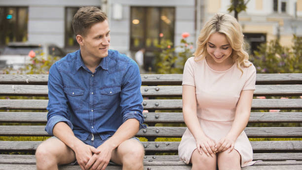 Shy blonde girl smiling, attractive guy flirting with beautiful woman on bench stock photo