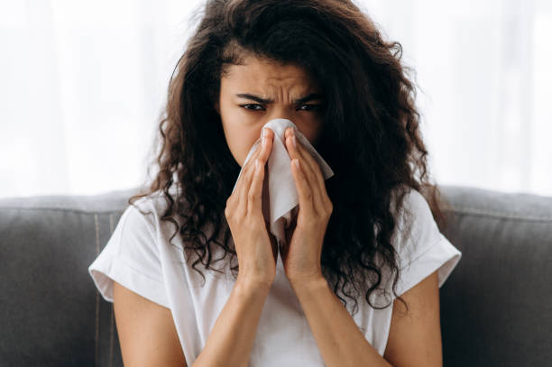 Sick african american girl sit on couch feel unhealthy, she suffering from rhinitis snuffles, having respiratory infection, get flue,  sitting at home at quarantine stock photo